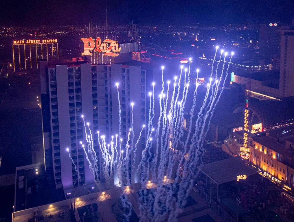 An aerial drone photo capturing a recent Plaza fireworks show. Several bright white fireworks light up the night sky with the Plaza neon sign visible behind the white light streaks in Downtown Las Vegas.