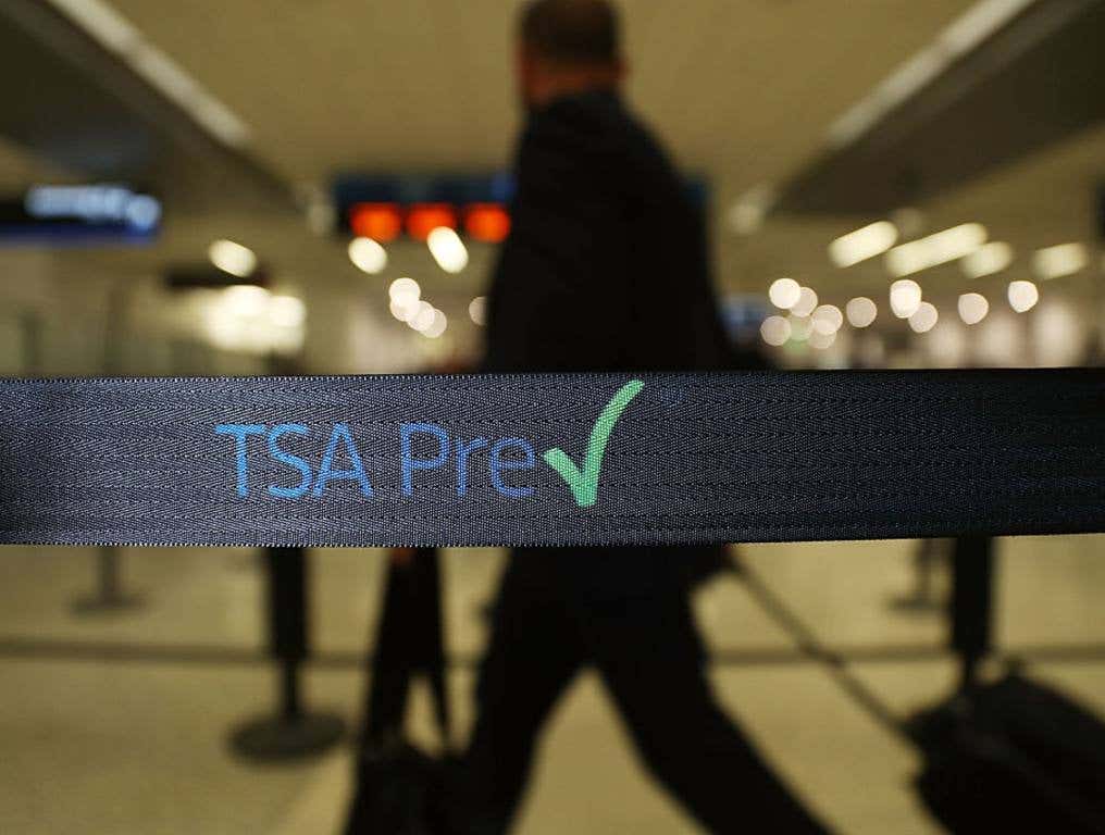 Photo of a male traveler walking through the TSA Precheck line at Miami International Airport. The focus of the photo is on the black TSA Precheck logo on black vinyl between two stanchions. The male traveler appears to be dressed in black and is rolling his carry-on suitcase behind him through the dimly lit airport terminal.