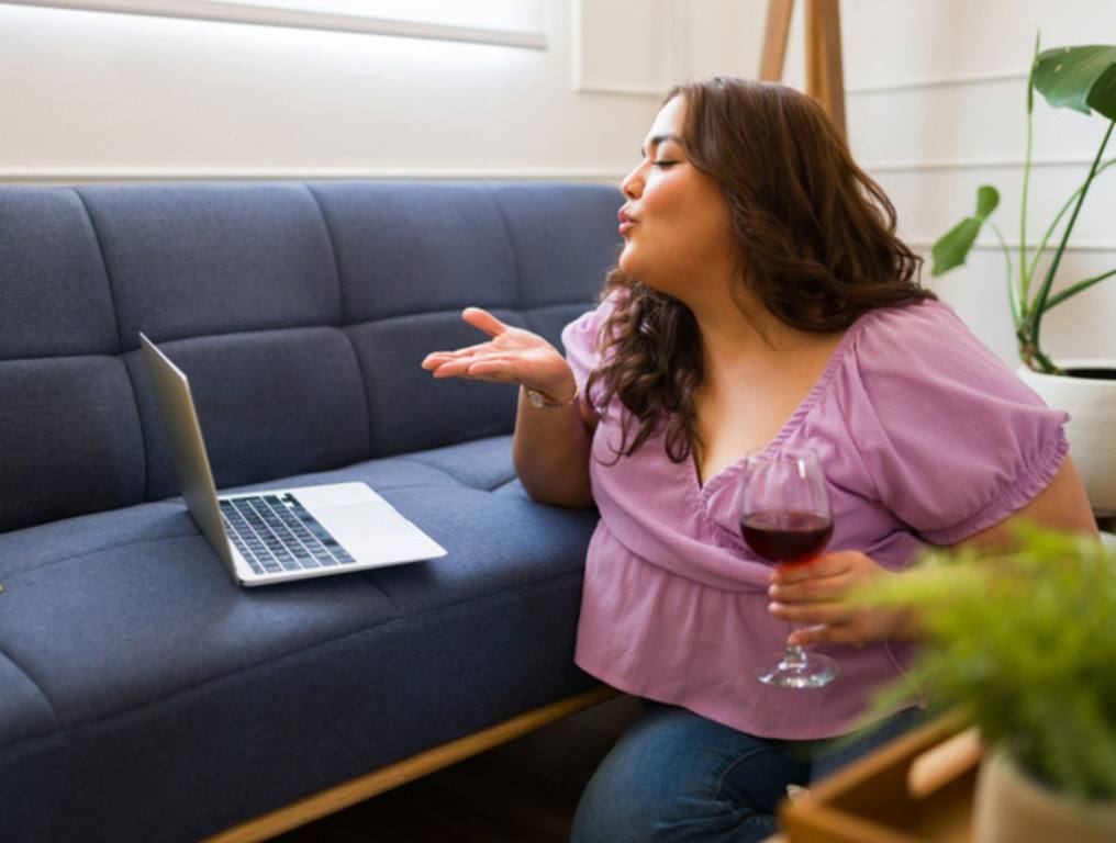 Romantic woman blowing a kiss to the screen to her long-distance boyfriend during an online video call with a glass of wine, virtual dates concept.