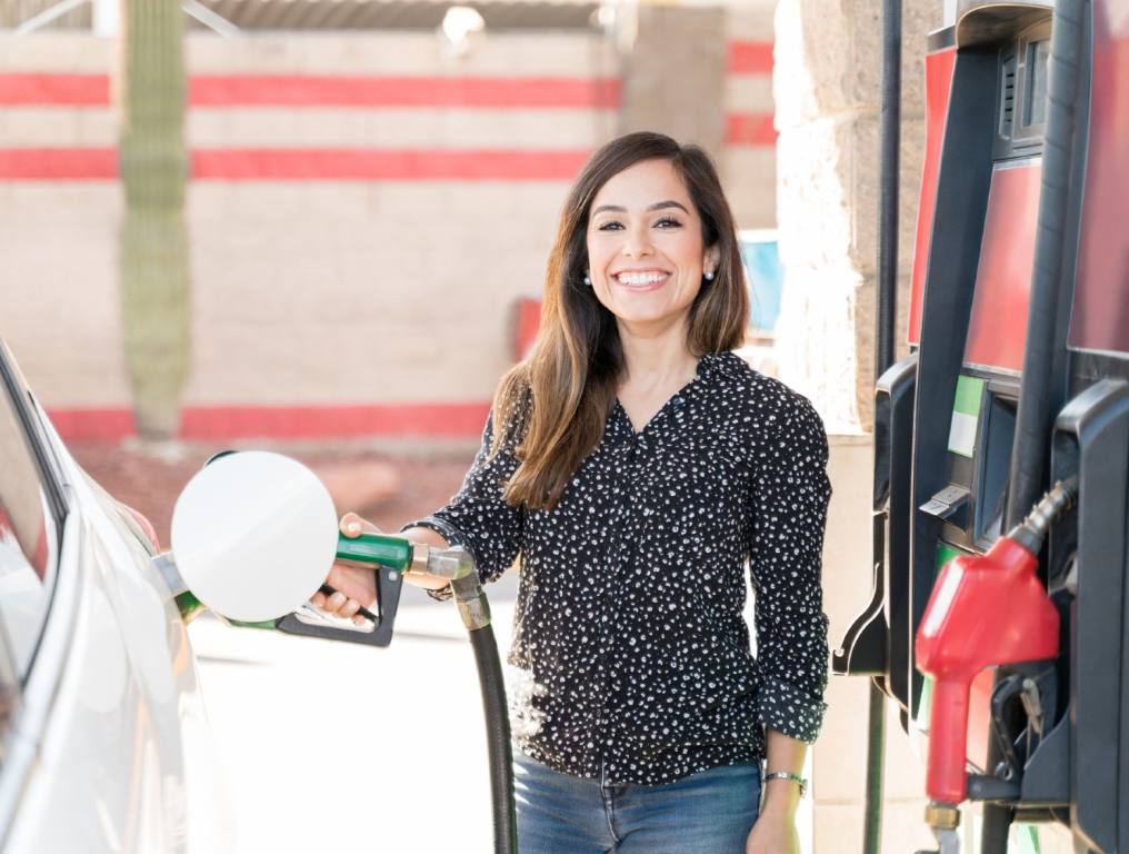 Photo depicting happiness when taking advantage of the Vegas Golden Knights free gas event. Photo of a young Caucasian woman in her late 20's who is smiling as she pumps gasoline into her white automobile. She is wearing a black long sleeved, button-down shirt with white polka dots all over it. She has chest-length brown hair and she is also wearing blue jeans. It appears to be a sunny day. The gasoline pumps are to her left. She is holding the nozzle with the green handle in her right hand.