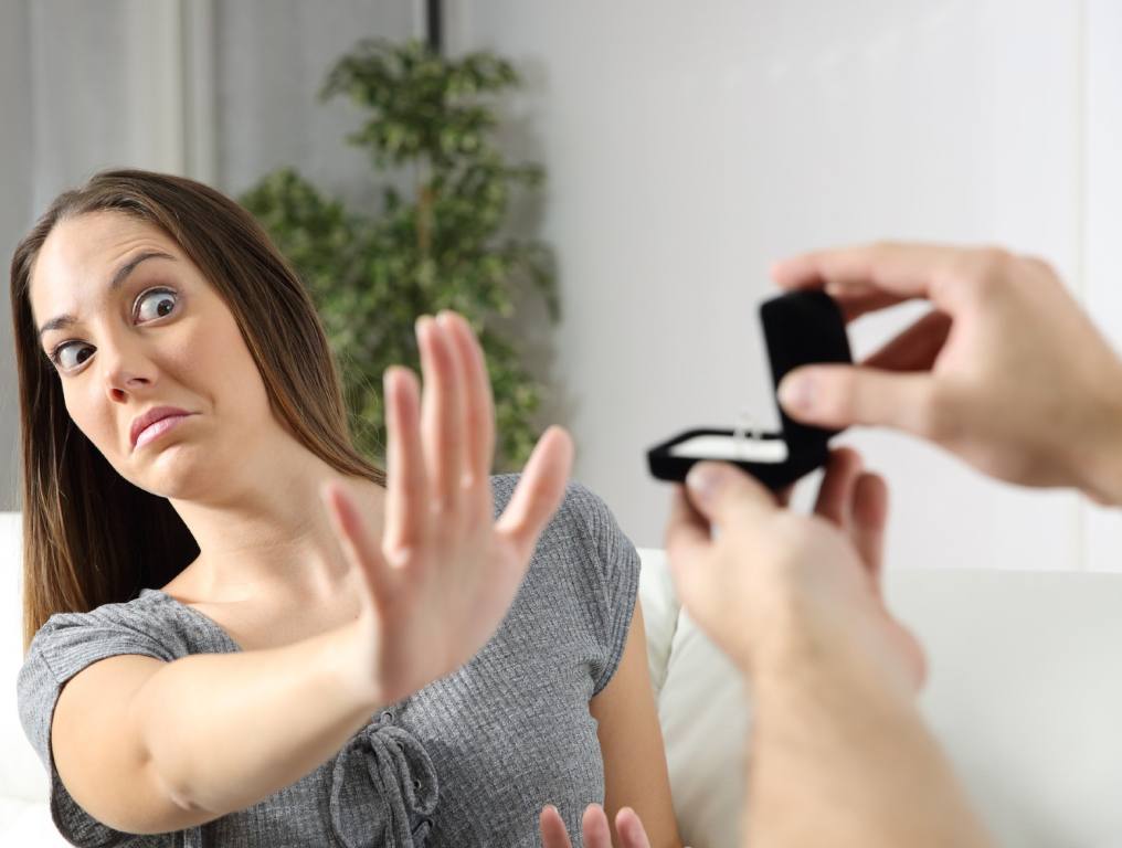 Marriage rates have seen a huge decline in Nevada since 2000. Photo of a woman who looks scared. She is holding up her right hand with her palm facing a person holding up a black velvet engagement ring jewelry box towards her. The woman has straight brown hair and she is wearing a gray t-shirt. It appears she is standing in her living room. There is a tall green plant in the corner of the room behind her.