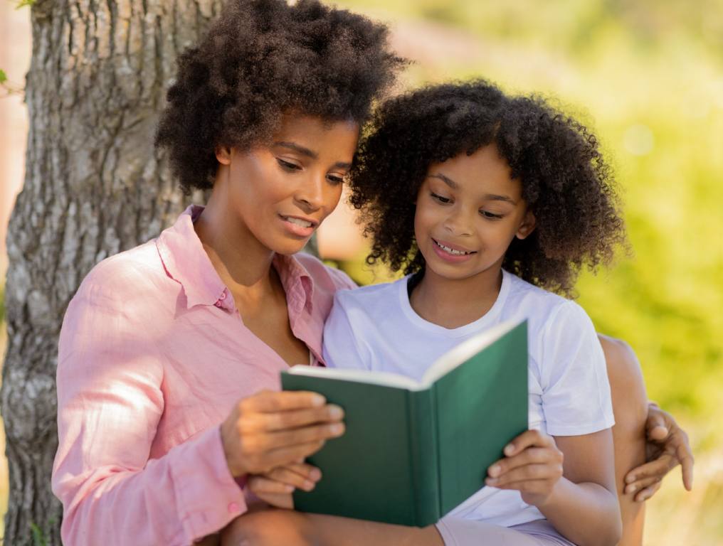 World Literacy Foundation is recruiting Youth Ambassadors to help fight global illiteracy. Photo of a high school or college female student helping a grade school student with their reading skills. The older young lady on the left is wearing a pink, button-down shirt which is open at the collar. The younger female student on the left is wearing a white t-shirt. The older student has her right hand on the right side of a green, hardcover book, and the younger student on the left has her left hand on the left side of that same book. They're both smiling on what appears to be a nice, sunny day. They are sitting up against a tree trunk in a park setting.