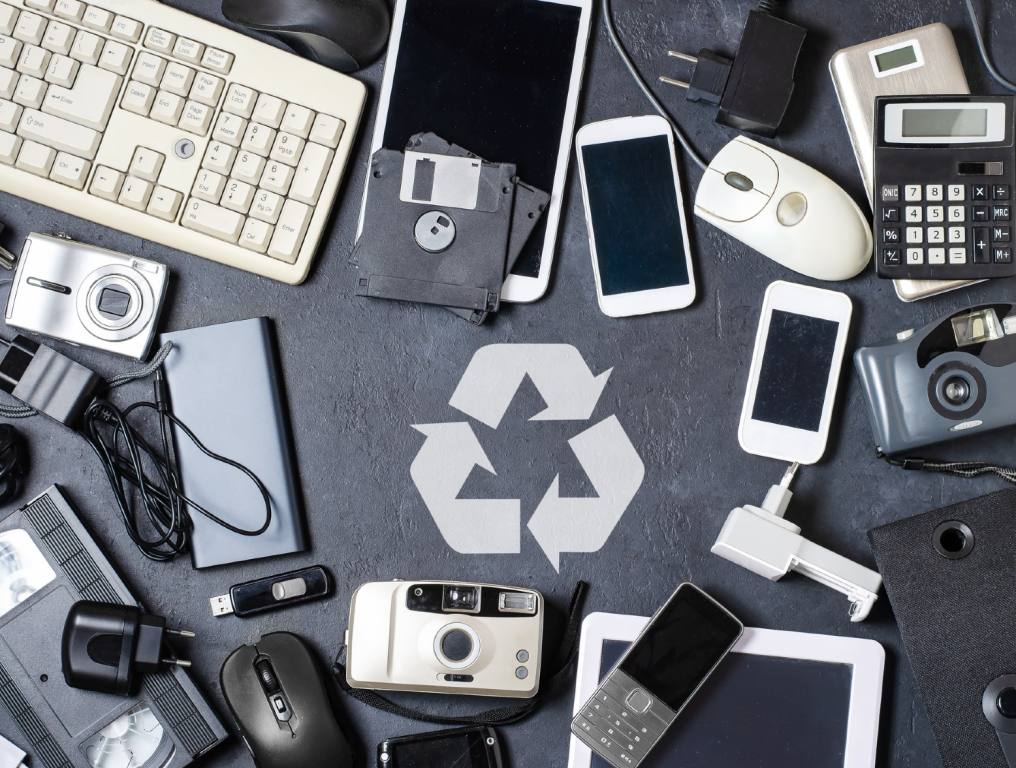 Here are some of the items you can bring to the Henderson Shines community clean-up for recycling. Black and white photo of outdated electronic equipment piled on a table or floor colored charcoal gray. There's a recycling symbol in the middle of all the equipment which includes mobile phones, cameras, a calculator, computer equipment, and videos.