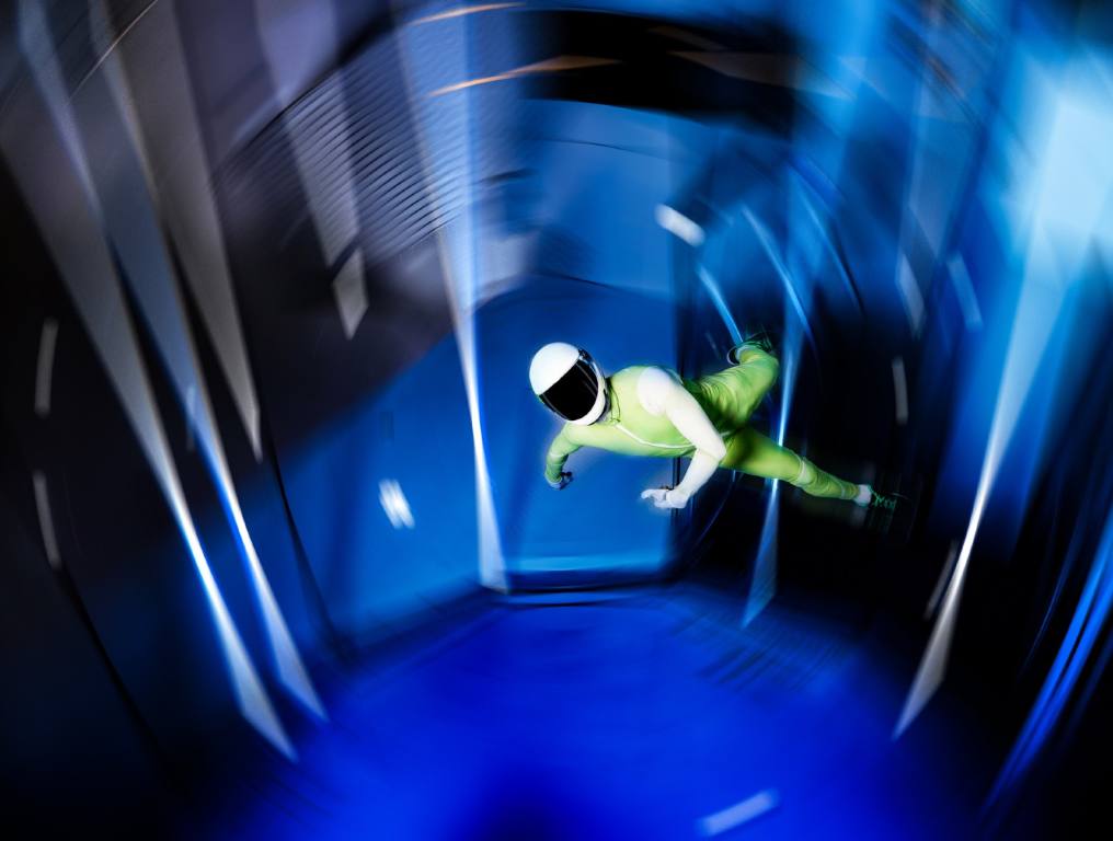 Crash n Burn on Fremont Street is opening Aero Vegas this summer 2024. Photo of a man doing stunts in an indoor wind tunnel. Man is wearing a bright green and white full body jumpsuit. He is also wearing a white skydiving helmet with a dark tinted shade covering his face. The photo is blurred to show circular movement while he performs his stunts in the wind tunnel.