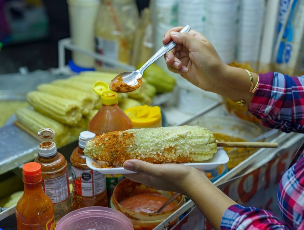 There are new restrictions and big fines in effect for unlicensed Clark County street vendors. Photo of a female street vendor preparing a serving of Mexican-style corn on the cob, called Elote. You can see the woman's hands, with sleeves of a colorful plaid shirt rolled up midway between her wrists and elbows. She is holding a metal tablespoon in her right hand as she seasons the Elote. There are corn cobs and other ingredients on her street vendor cart or table in the background.