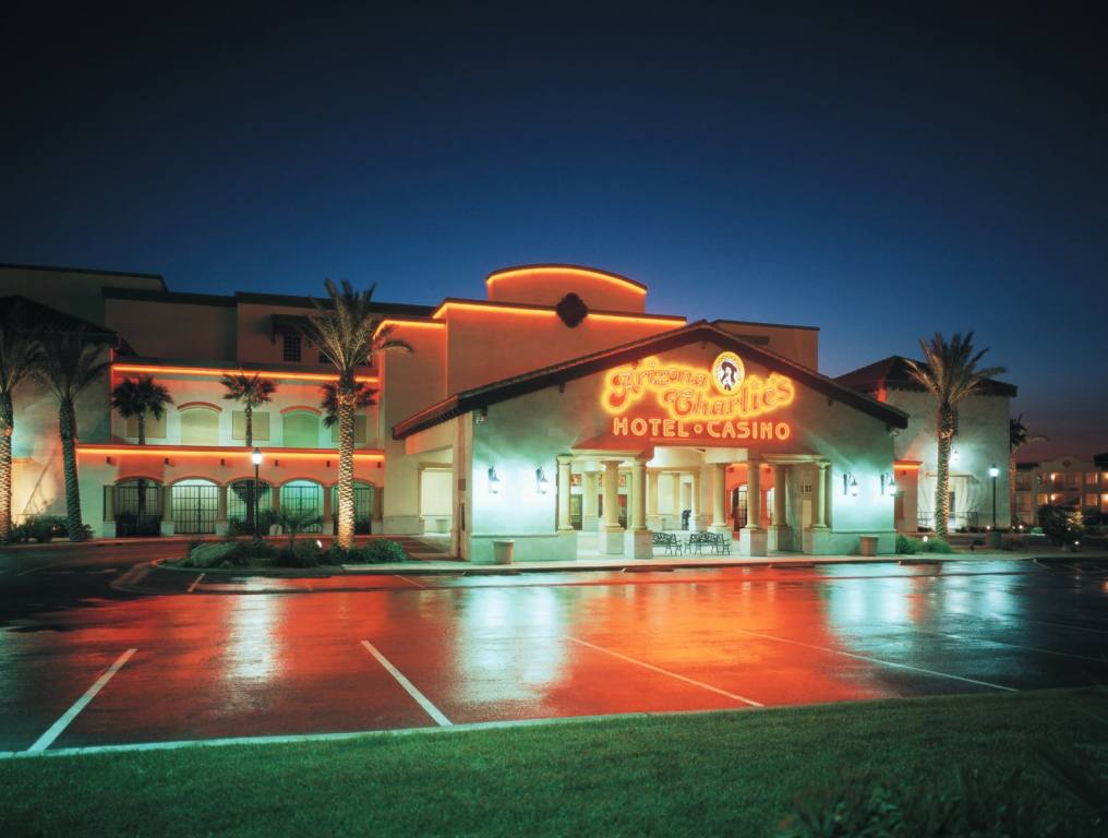 Exterior facade of Arizona Charlie's Boulder location. It is nighttime and the lights are on, showing orange lettering. The parking lot is empty and the pavement is wet, reflecting the lights.