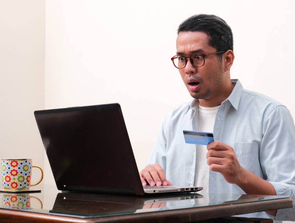 Many Las Vegas valley residents are losing thousands of dollars in the latest debit card scam. Photo of a man in his early 30's with a look of shock and surprise on his face while he looks at his laptop computer. He is holding a credit or debit card in his right hand while staring at his laptop screen, which is sitting on a glass desktop. He has a coffee cup to the right of his laptop. He is wearing a light blue button-down shirt with the sleeves rolled up to the middle of his forearms and the front of the shirt unbuttoned. He is wearing a white t-shirt under the button-down. He is sitting in a room or office with white walls.