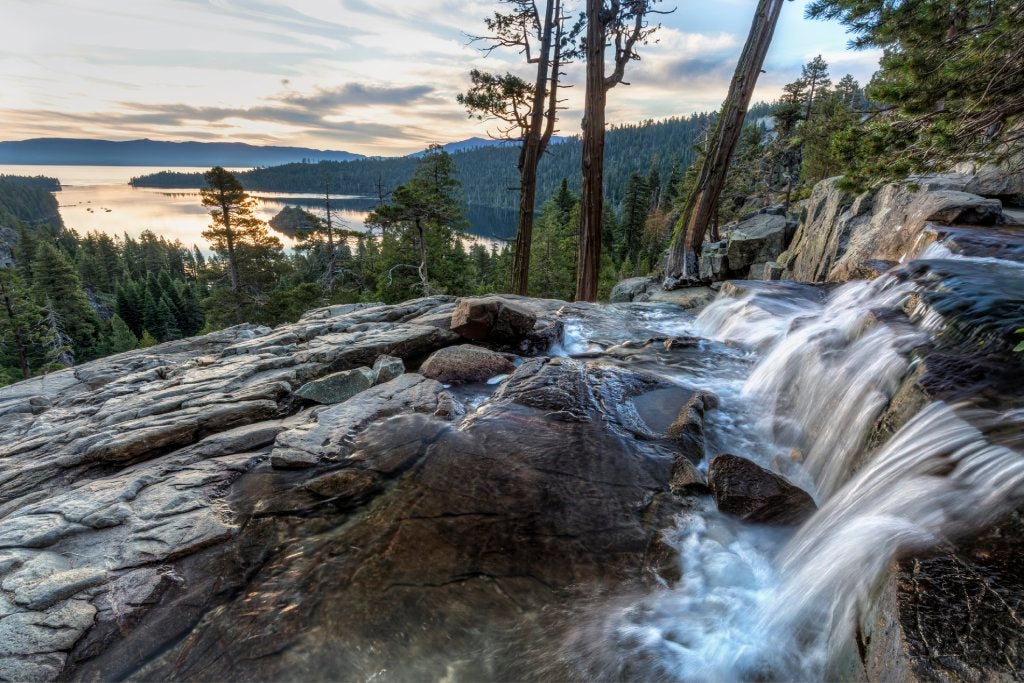 Distant view of a lake with a waterfall over rocks in the foreground and pine trees in the midground.