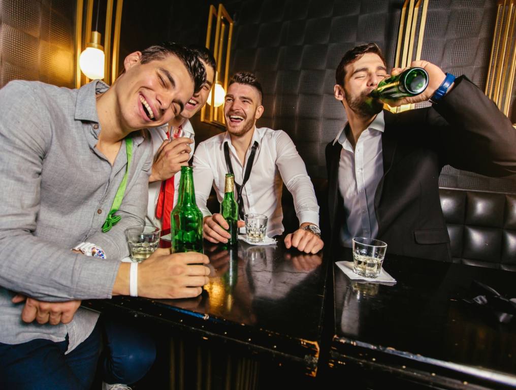 Four friends at a Vegas Bachelor Party are drinking in a nightclub. They're all wearing button up shirts and loosened ties.