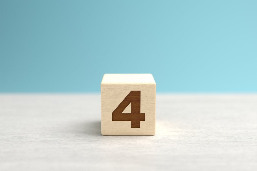 A wooden toy cube with the number four on it sits on a gray surface in front of a blue backdrop.