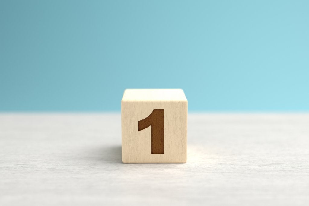 A wooden toy cube with the number one on it sits on a gray surface in front of a blue backdrop.