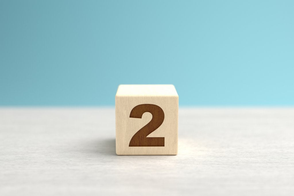 A wooden toy cube with the number two on it sits on a gray surface in front of a blue backdrop.