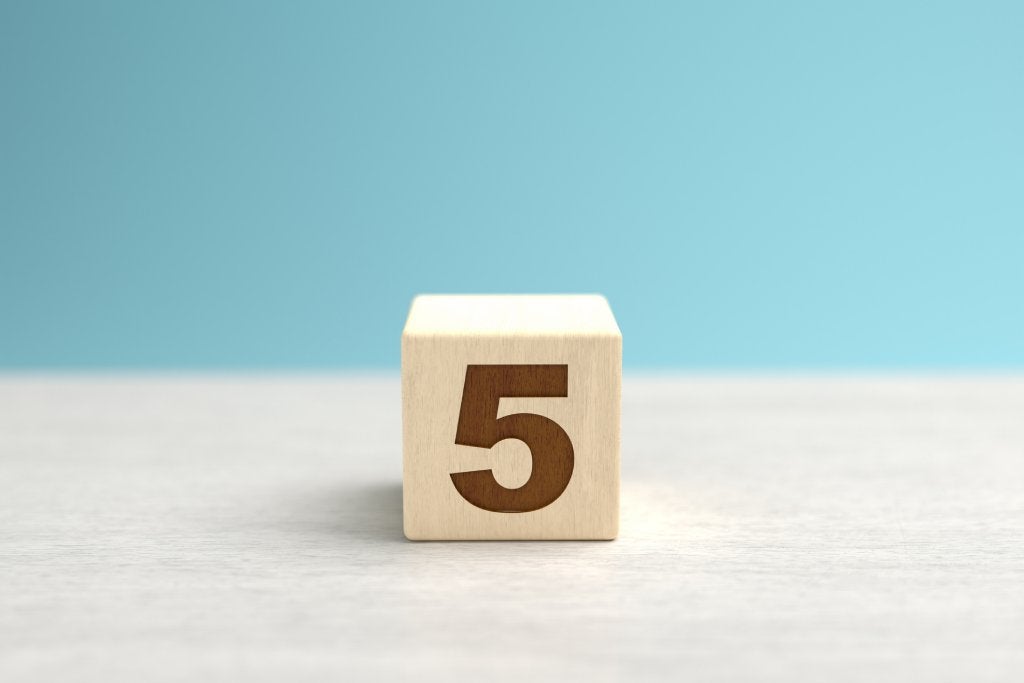 A wooden toy cube with the number five on it sits on a gray surface in front of a blue backdrop.