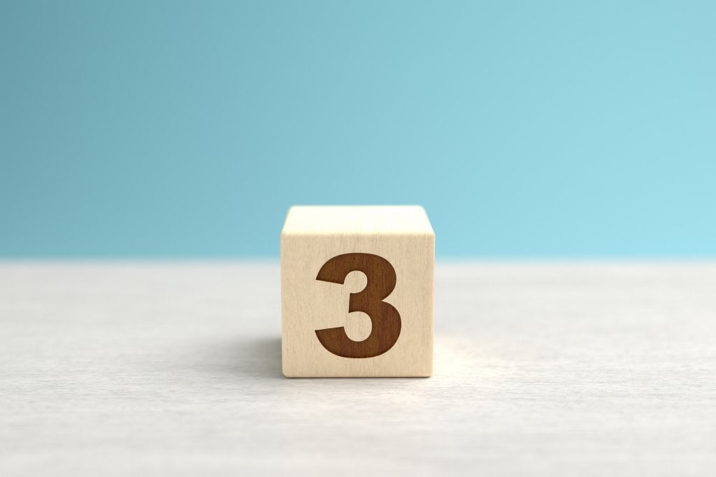 A wooden toy cube with the number three on it sits on a gray surface in front of a blue backdrop.