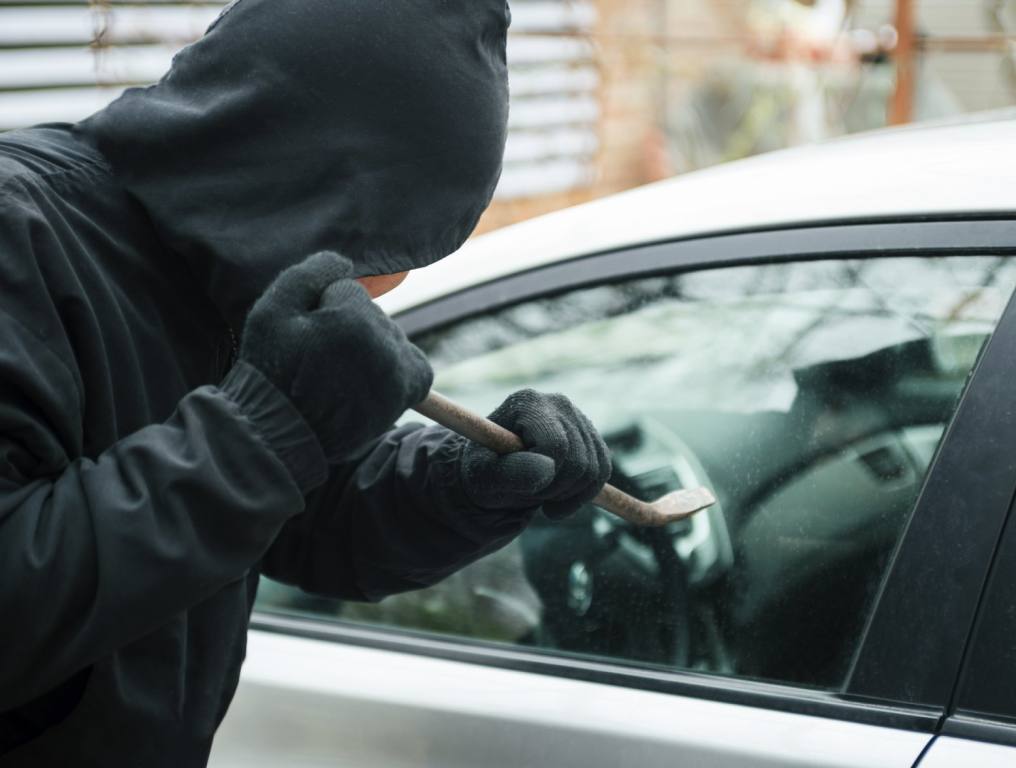 A man in a black hoodie is using a crowbar to break into a silver car. Concept of Nevada residents car theft.