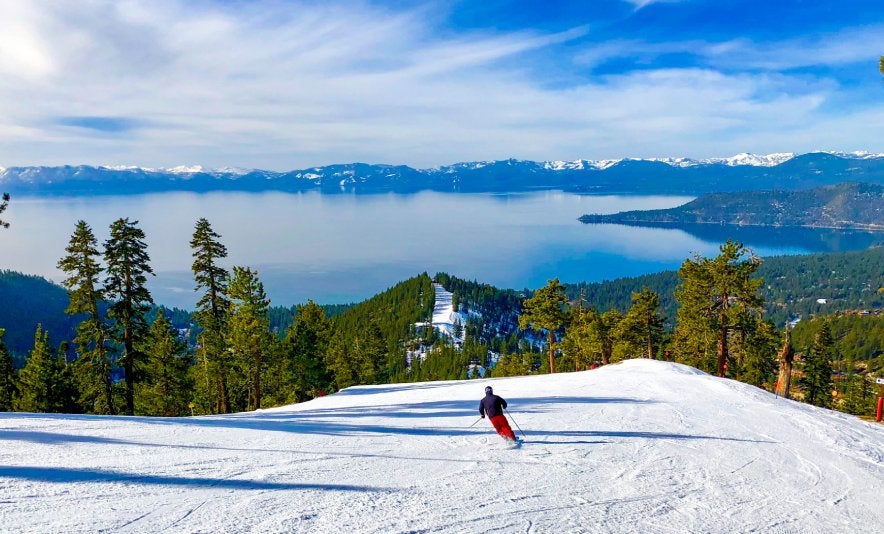 A lone skier on a snow-covered mountain overlooking a lake and pine trees.