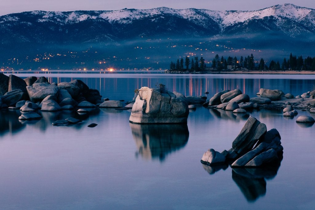 A very still lake reflects large rocks in the foreground and lights and mountains on the shore across the lake . It is near dusk.