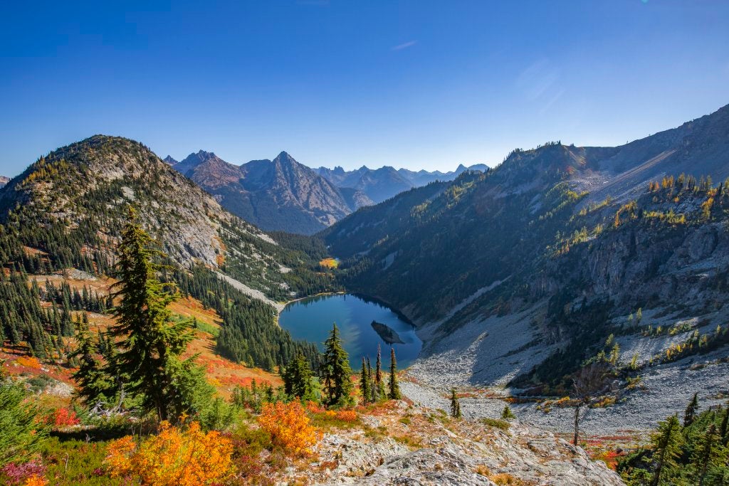 Aerial view of a lake surrounded by tall mountains covered with pine trees under a blue sky.