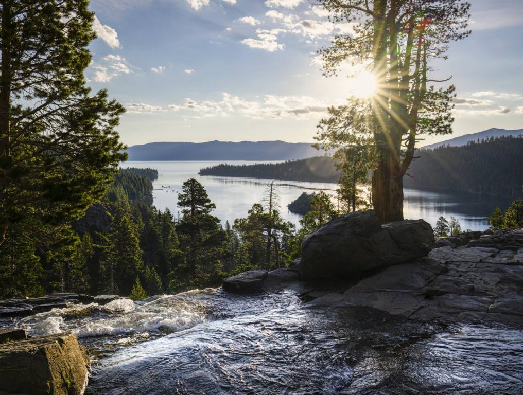 View from a mountain waterfall overlooks a lake and more mountains through the branches of pine trees. A sunrise is seen in the distance and the sky is yellow and blue. Lake Tahoe