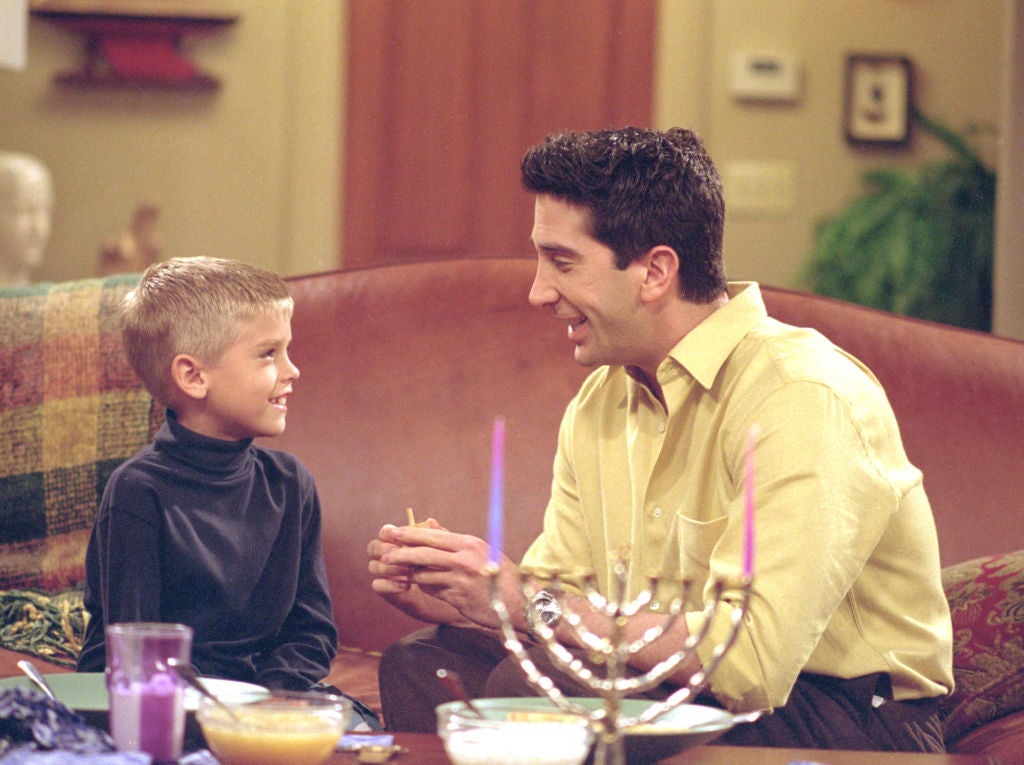 A blonde boy sits on a couch on a television set across from a dark-haired man playing his father. They are talking while a menorah stands on the coffee table.