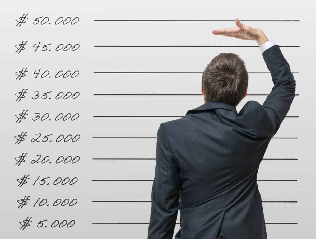 Enterprise in Southwest Las Vegas is now the neighborhood with the highest median income. Photo shows the back of a man, wearing a dark gray business suit. He is standing in front of a whiteboard with salaries listed on the left from $5,000 dollars to $50,000 dollars. He is measuring his height against the lines on the whiteboard as if measuring his salary versus average wages.