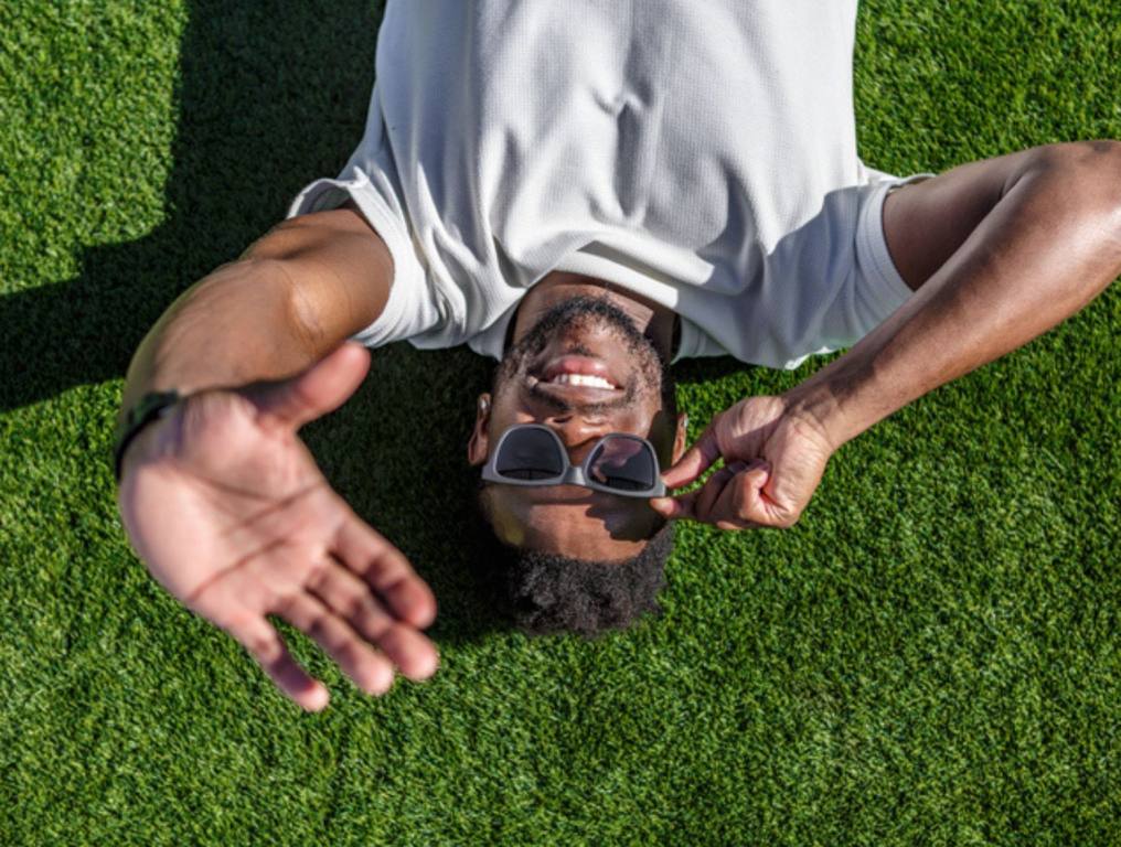 An African-American man in his 30s wearing sunglasses lies sprawled on the grass of a well-maintained public park.