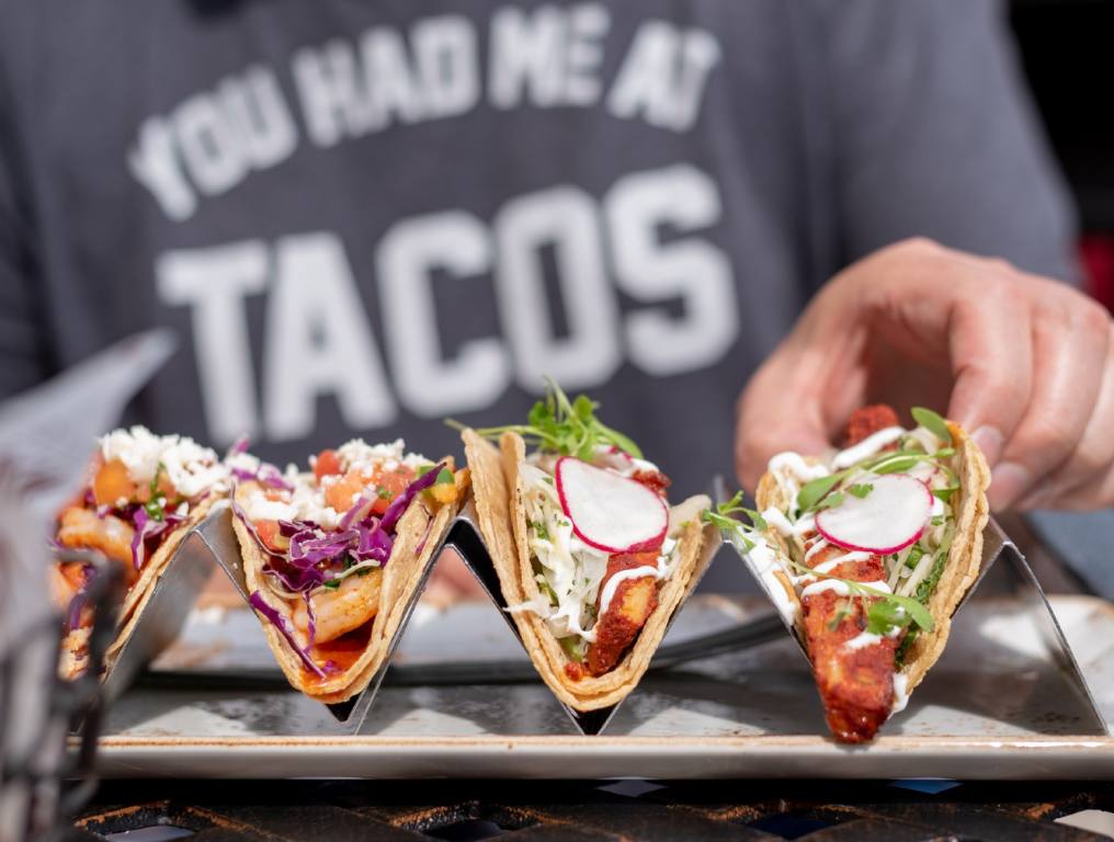 Las Vegas is named best U.S. city for taco lovers! Photo of a male chef preparing four different varieties of tacos, and placing the final one in the fourth slot on the far right of the taco holder. You can only see the chef's left hand in focus. In the background behind the tacos, slightly out of focus, you can see the chef's torso. He is wearing a gray t-shirt with white block letters reading "You had me at TACOS."