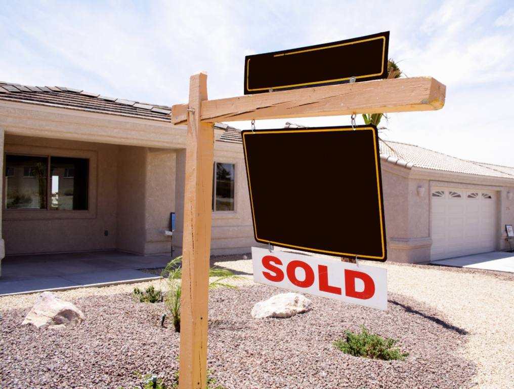 Nevada houses are expected to be the second most expensive in the U.S. by 2030. Photo shows a brown realtor sign on an unpainted wooden post. Sign reads "Sold" in red letters on a white background. The tan stucco, one-story house has sparse desert landscaping out front.