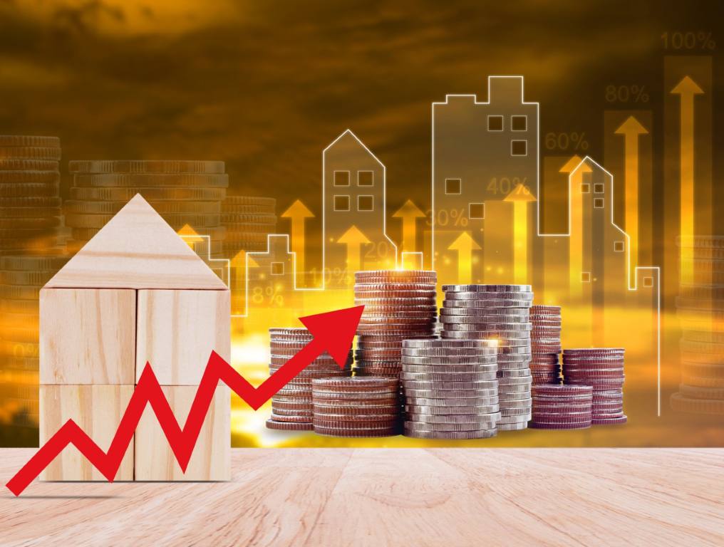 Rent increases outpacing wages in Las Vegas, and the majority of U.S. cities over the past 5 years since the COVID-19 pandemic. 3-D rendering shows an unpainted toy wood house on the left. A jagged rising red arrow is superimposed in the foreground of the image. There are 8 stacks of quarters, of varying heights, on the wooden table to the right of the wood toy house. In the background, opaque images of apartment buildings in varying shades of yellow and gold can be seen. There are rising yellow arrows superimposed over those apartment building images.