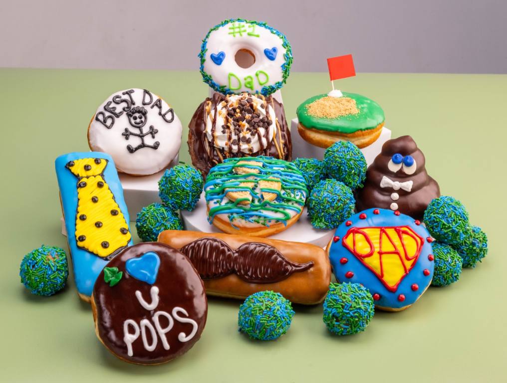 A collection of Father's Day donuts sitting on a green table includes pastries decorated with ties, mustaches and other dad-related images.