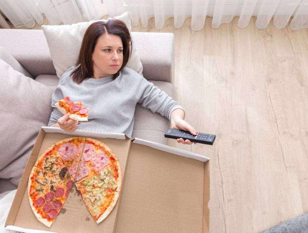 QA woman being lazy on the couch watchin tv and eating pizza.