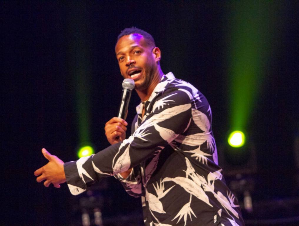 Comedian Marlon Wayans is performing on stage wearing a black and white shirt and holding a microphone. A 2024 Marlon Wayans tour is coming to Las Vegas.