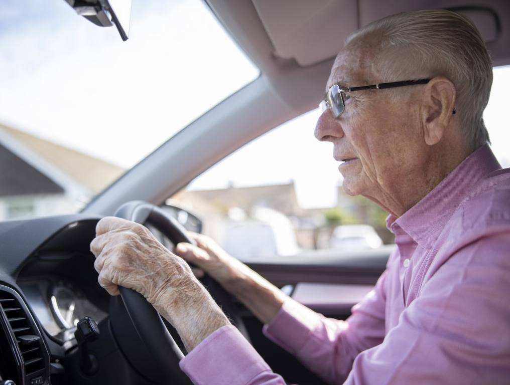 An elderly man holds tight to his steering wheel as he drives wearing glasses and a pink button-up shirt. Concept of safest drivers on the road.