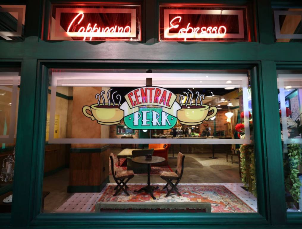 A replica of the Central Perk Cafe from Friends show the window with the cafe logo and the cafe itself seen through the window in the background. Concept: The Friends Experience