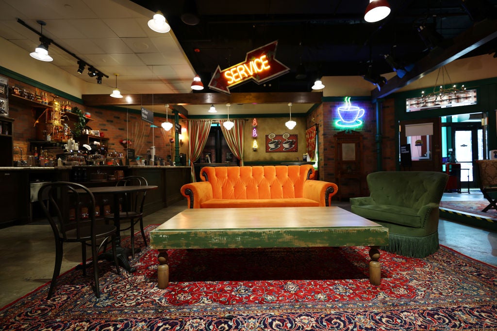 An orange sofa is under a spotlight on a tv show set surrounded by cafe furniture and accessories.