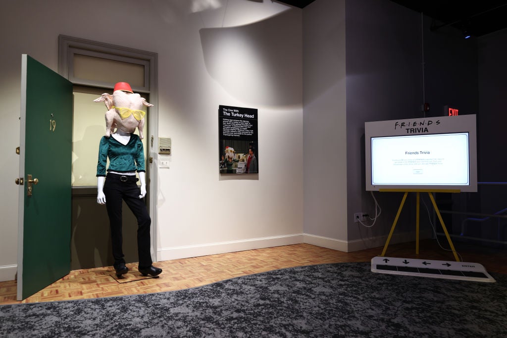 A mannequin dressed in black jeans and a green shirt is positioned in the entryway of a door with a turkey, hat and sunglasses on its head.