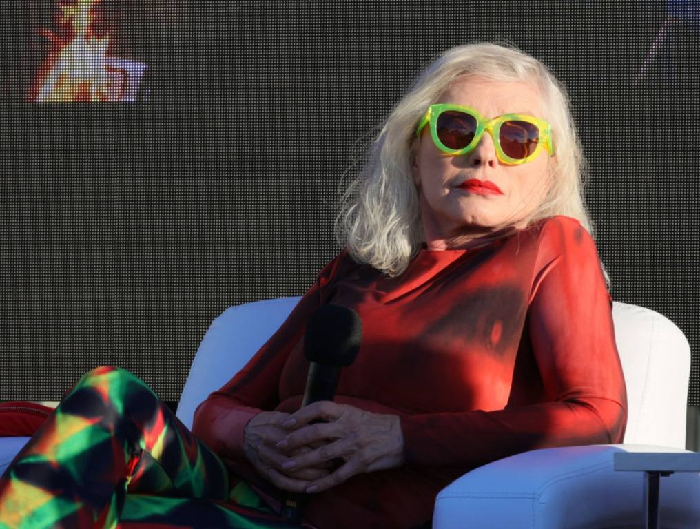 Singer and actress Debbie Harry sits in a chair leaning on her elbow. She is wearing neon green sunglasses and a red shirt. Debbie Harry acting concept.