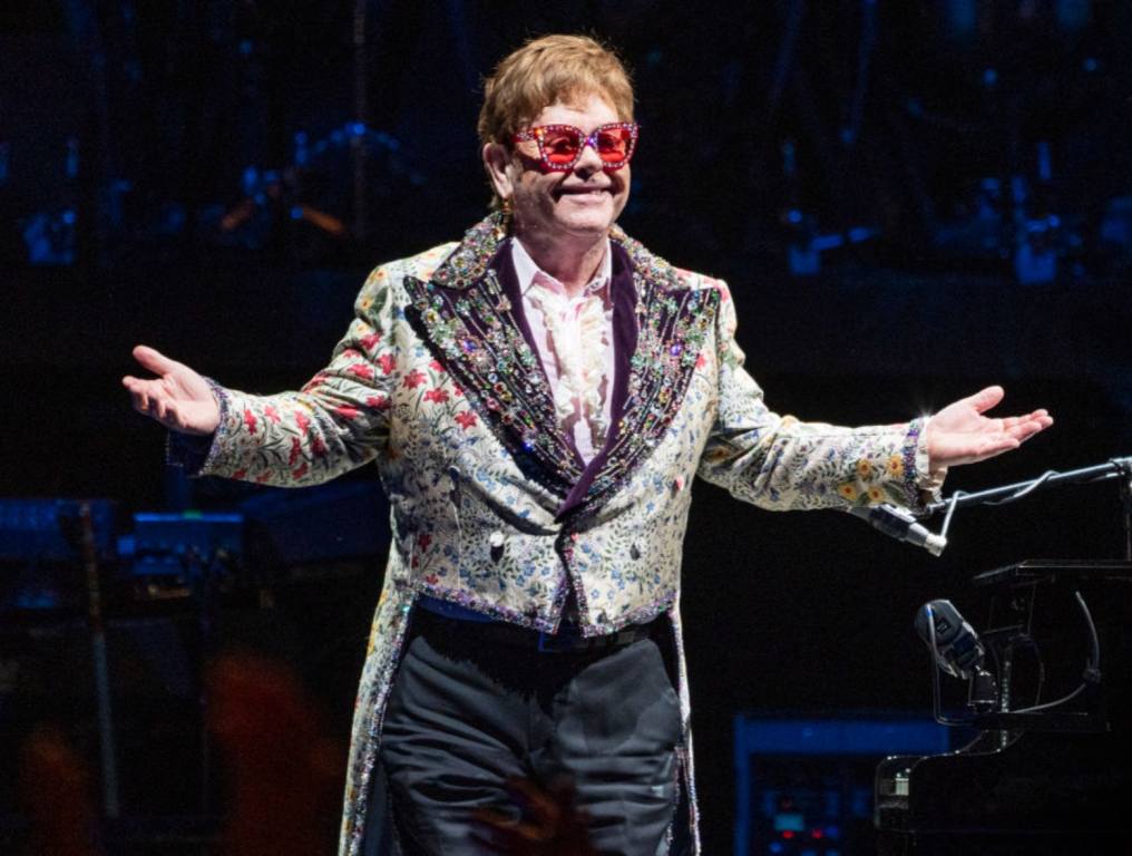 Performer Elton John on stage smiling at his audience in a white jacket with tails and pink sunglasses. Concept of Elton John's outfits sold at auction.