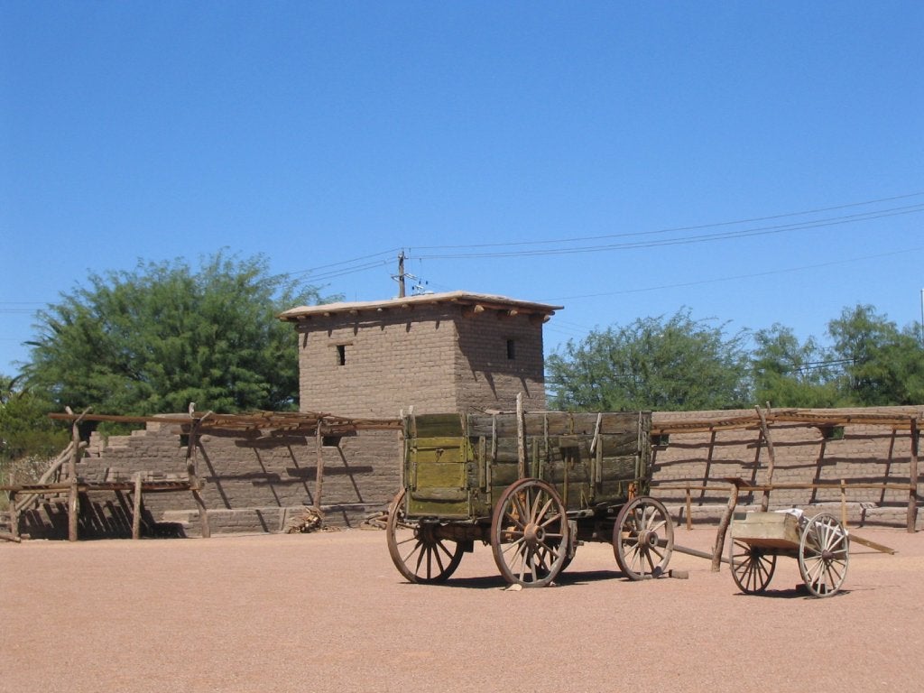 An old square building sits on a dirt lot with a wooden fence and old wagon.