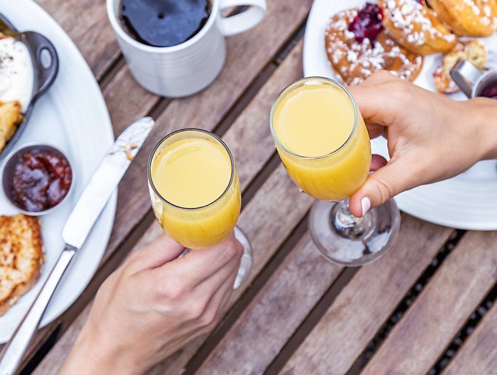 Close-up of two hands holding champagne glasses filled with mimosas. They are about to clink the glasses over a table of brunch food. Concept of Vegas brunches.