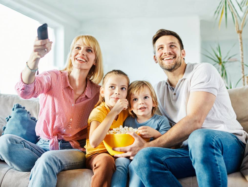 A family of four sits on a couch eating popcorn and watching a movie. Mom has the remote and dad is leaning over the young son and daughter on the couch. Concept of Nevada's favorite musicals.