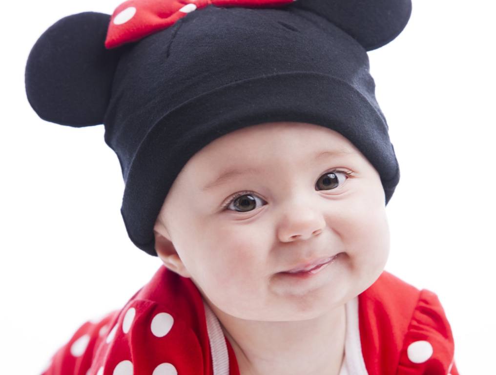 An infant girl looks at the camera against a white backdrop. She is wearing a red outfit with white polka dots and a beanie with mouse ears and a matching bow. Concept: Disney baby names