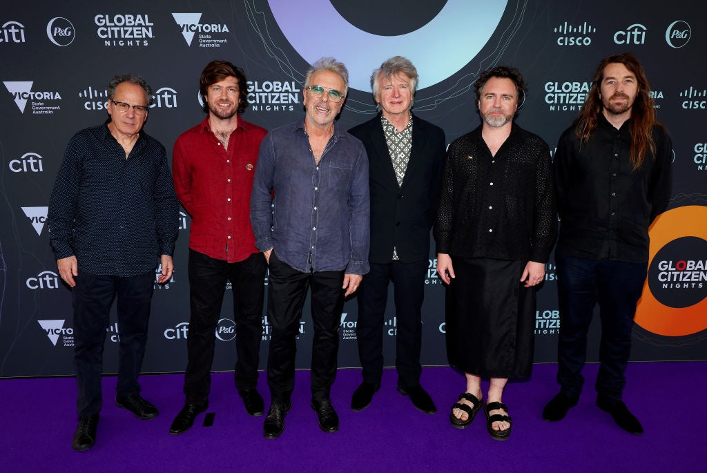 Six members of Crowded House stand in a row and pose in front of a banner.