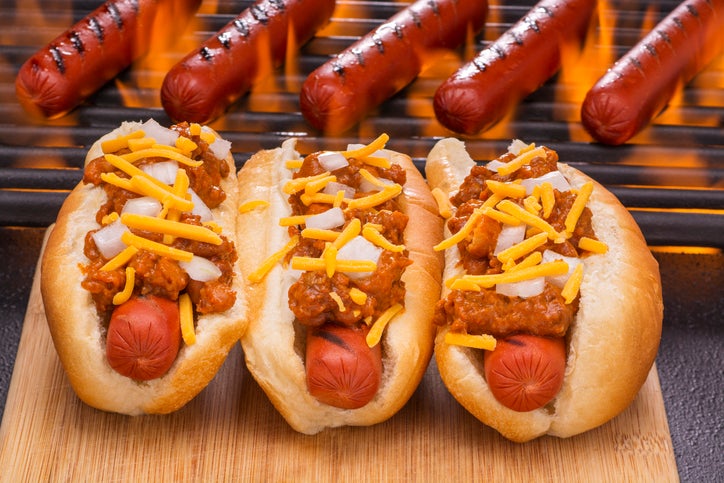 chili hot dogs with cheddar cheese and diced onions next to hot dogs grilling on a flaming hot barbecue. the best chili dogs ever