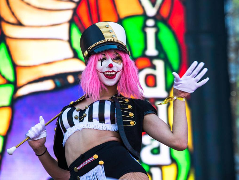 A young woman with pink hair and clown makeup smiles in a costume in front of a bright lit backdrop.