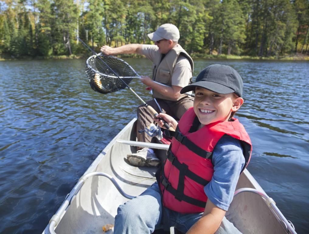 A young boy in a red life jacket smiles with a fishing pole in his hand as a man next to him in a canoe pulls a fish from a net. Concept: Nevada Free Fishing Day