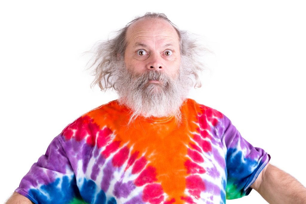 A balding older man with long white hair on the sides and white beard is wearing a tie-dyed t-shirt looking surprised.