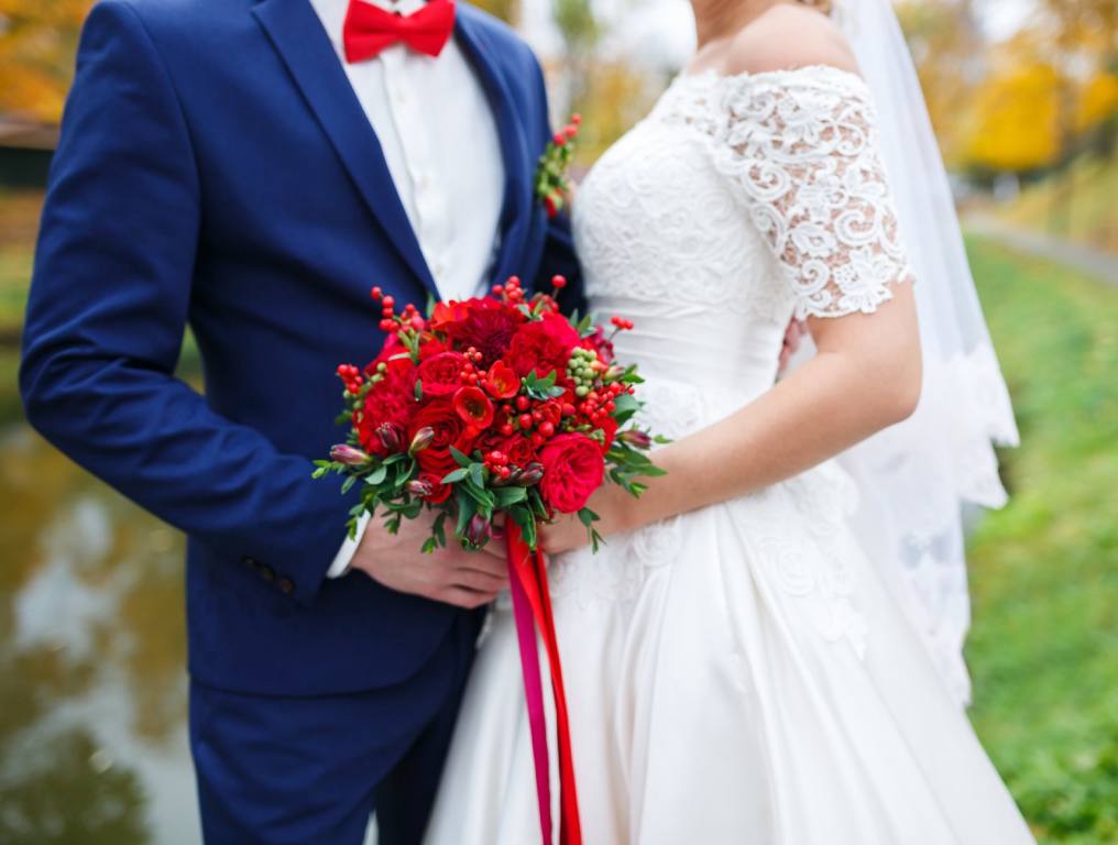 Torso shot of a couple getting married. The groom on the left is wearing a blue suit with red bowtie and the bride on the right is wearing a lacy white dress and holding a red bouquet. They are facing each other. Concept of patriotic wedding at The Little Vegas Chapel.