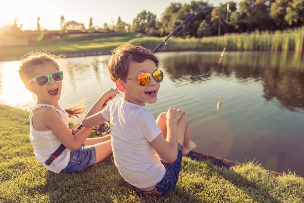 A boy and a girl sit on a grassy shore of a pond wearing sunglasses and holding fishing poles happily.