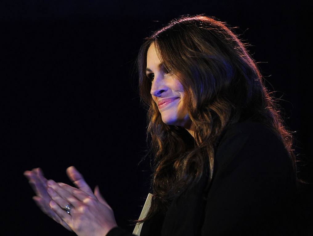 Julia Roberts is one movie star featured in hilarious movie scenes singing a big hit in the romantic comedy "Pretty Woman." A photo of actress Julia Roberts' head and shoulders in a darkened auditorium. She is applauding. She is wearing her dark brown hair down to her shoulders. She is wearing a black sweater and a diamond wedding ring.